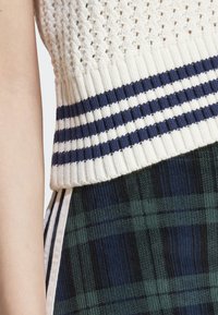 Top de malha branca com riscas em azul-marinho na bainha, combinado com uma saia xadrez em verde escuro e preto. Materiais texturizados aumentam o interesse visual.