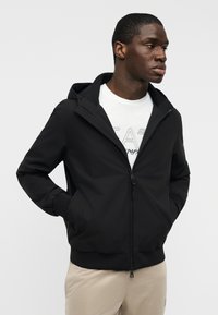 Μαύρο τζάκετ με φερμουάρ και κουκούλα, με ελαστικά μανσέτα και τσέπες. Φορεμένο πάνω από λευκό πουκάμισο με γραφική εκτύπωση. Λείο, ελαφρύ ύφασμα.