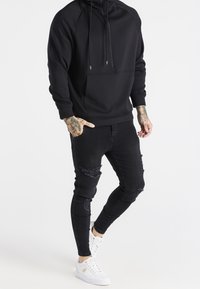 Černá mikina s kapsou na zip, v kombinaci s potrhanými černými skinny džíny a bílými teniskami s zlatými akcenty.
