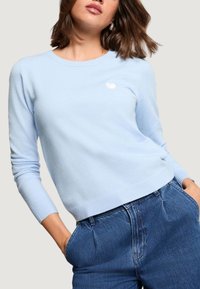 Jasnoniebieski sweter z dzianiny z okrągłym dekoltem i długimi rękawami; na piersi znajduje się małe białe logo w kształcie serca, a mankiety są ściągane.