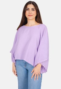 Lavendelfarbenes, locker sitzendes Oberteil aus strukturiertem Stoff mit weiten Ärmeln, das Spitzenakzente am Saum aufweist. Kombiniert mit hellblauen Jeans.