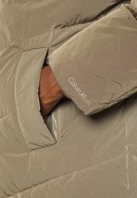 Chaqueta acolchada beige con un acabado texturizado, que presenta un bolsillo y un puño con "Calvin Klein" bordado en blanco.