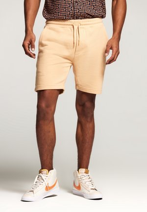 Hellbeige Baumwollshorts mit einem elastischen Bund und Kordelzug. Sie haben eine lockere Passform mit umgeschlagenen Säumen. Kombiniert mit weißen Sneakers.