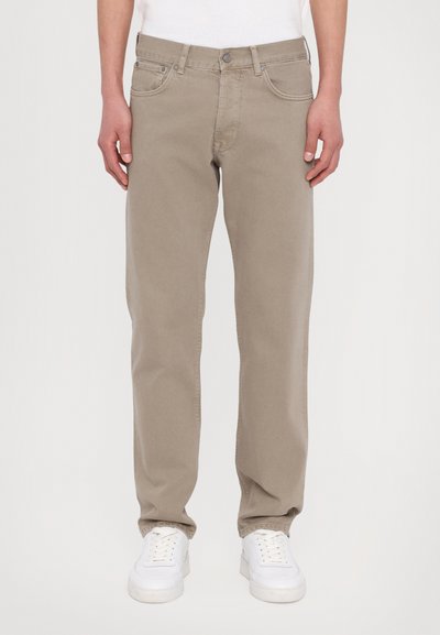 Jean beige tapered avec cinq poches, fermeture à bouton et passants de ceinture, associés à des baskets blanches. Le tissu semble lisse et uniforme.