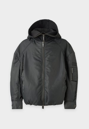 Un mannequin masculin se tient devant un fond blanc de studio. Il porte une casquette beige, un blouson bomber noir à capuche, un pantalon cargo gris ample et des baskets foncées.