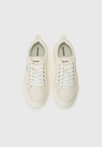 White canvas sneakers with a textured finish, featuring flat white laces, a padded collar, and branding on the tongue and side.