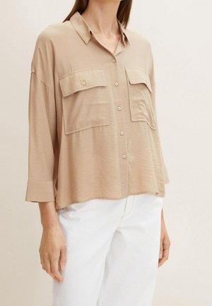 Femme portant une chemise beige ample à boutons avec deux grandes poches à la poitrine et un pantalon blanc ; sa tête n'est pas visible dans le cadre.