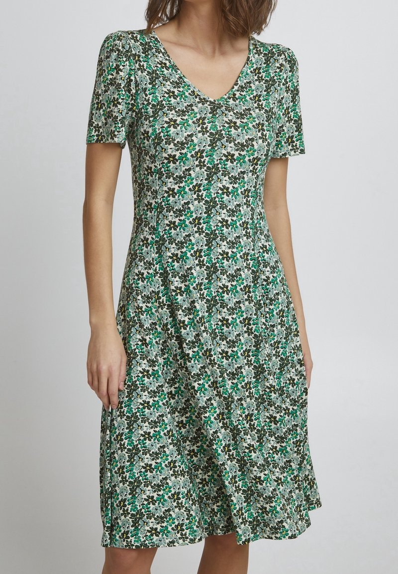 Robe florale avec un décolleté en V et des manches courtes. Motif floral vert, noir et jaune sur fond clair. Jupe évasée avec coutures latérales.