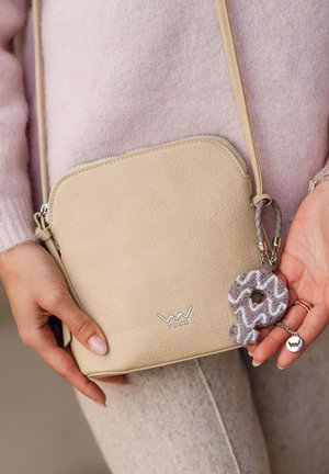 Béžová kožená kabelka přes rameno s kulatým tvarem, stříbrnými kovovými doplňky a texturovaným povrchem. Připojený je šedý přívěsek a klíčenka s potiskem.