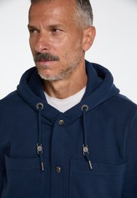 Sweat-shirt à capuche bleu marine avec deux poches avant, boutons-pression et cordons de serrage. Matière douce avec une texture lisse.