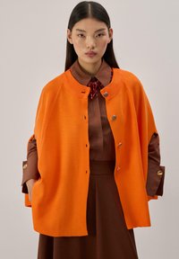 Cardigan orange en maille avec un col rond et des détails de boutons argentés, porté sur une chemise marron à col. Le tissu a une texture lisse.