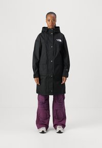 Parka noire imperméable avec une capuche, fermeture par boutons-pression à l'avant, deux grandes poches et un logo sur le côté gauche de la poitrine. Portée par-dessus un pantalon violet ample.
