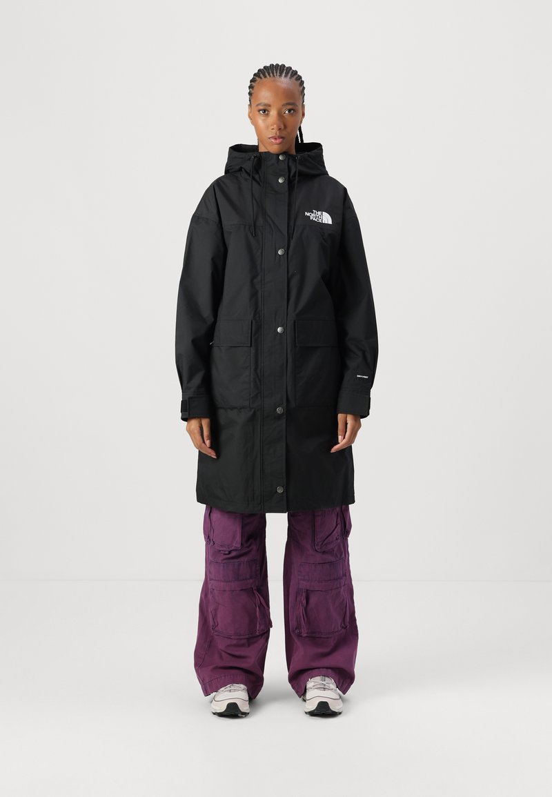 Parka noire imperméable avec une capuche, fermeture par boutons-pression à l'avant, deux grandes poches et un logo sur le côté gauche de la poitrine. Portée par-dessus un pantalon violet ample.