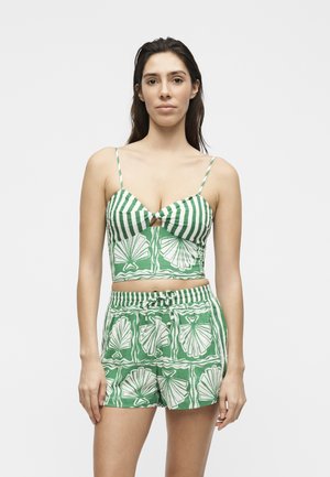 Γυναίκα με πράσινο και λευκό ριγέ κοντό τοπ και ταιριαστά σορτς με σχέδια από κοχύλια, στέκεται μπροστά σε απλό φόντο.