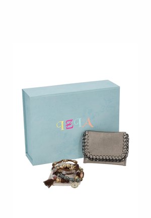 Una cartera gris metalizada con detalles de cadena y varias pulseras de cuentas superpuestas están delante de una caja de regalo azul claro.