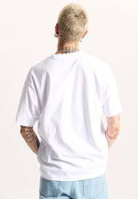 Camiseta de algodón blanca de manga corta, con cuello redondo y corte relajado; textura suave; tatuajes visibles en los antebrazos; jeans de mezclilla azul claro.