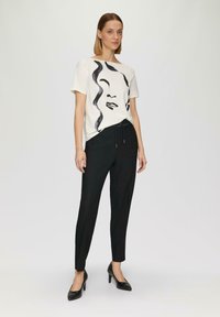 Witte korte mouwen shirt met een zwart abstract gezicht ontwerp, gecombineerd met zwarte tapered broek en zwarte hakken. Eenvoudige, moderne stijl.