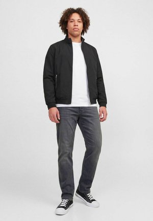 Jeune homme aux cheveux bouclés portant une veste noire, un t-shirt blanc, un jean gris et des baskets noires, debout devant un fond uni.