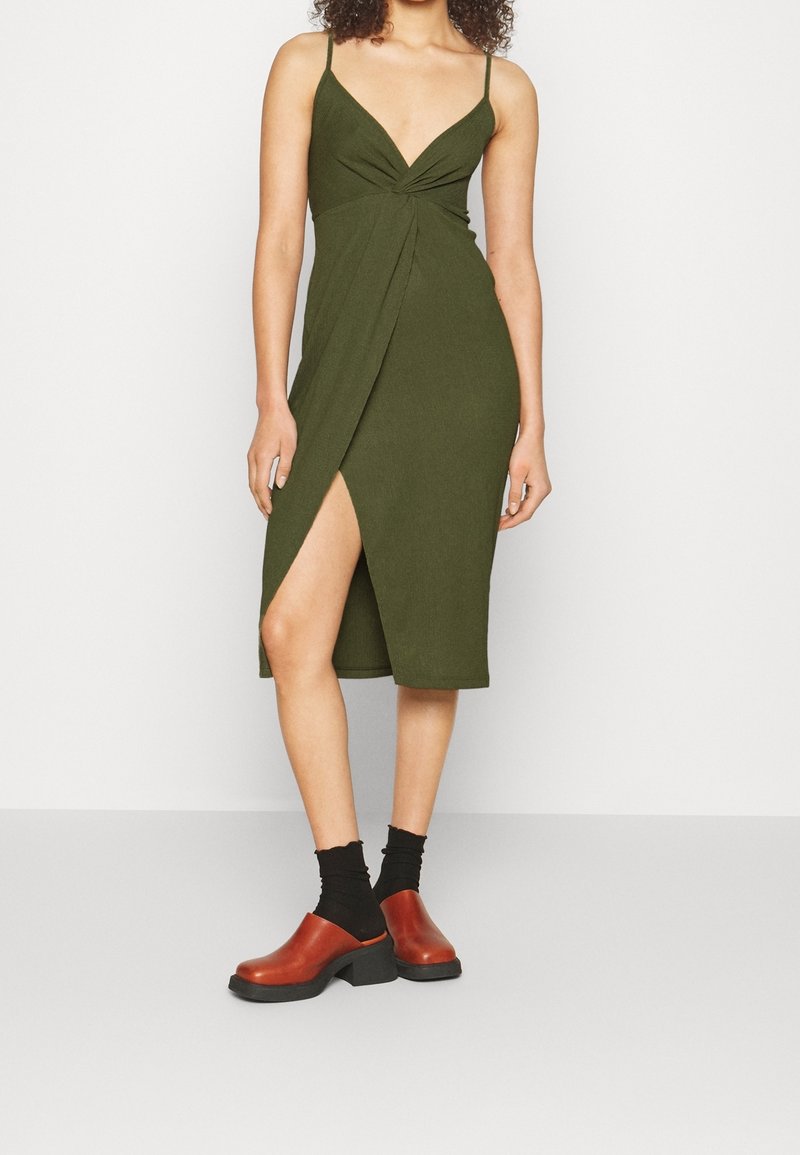 Femme portant une robe midi sans manches vert olive avec une fente à l'avant, des chaussettes noires et des sabots marron à talons sur fond uni.