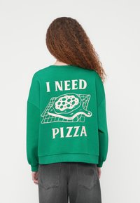 Πράσινη φούτερ μπλούζα με χαλαρή εφαρμογή, που έχει ένα μεγάλο γραφικό με ένα πίτσα σε δίσκο και το κείμενο «ΧΡΕΙΑΖΟΜΑΙ ΠΙΤΣΑ» σε κρεμ χρώμα.