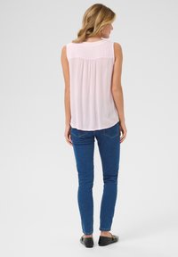 Jasnoróżowa bezrękawnikowa bluzka z plisowanym tyłem, zestawiona z niebieskimi skinny jeansami i czarnymi balerinami, prezentowana od tyłu.