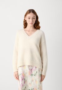 Suéter crema de cuello en V con una textura suave y difusa, y ribete acanalado, llevado sobre una falda con un estampado floral.