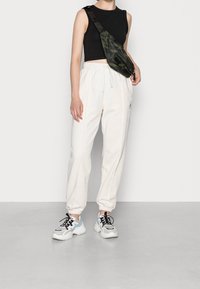 Person med svart ärmlös crop top, vita joggerbyxor med dragsko, kraftiga vita sneakers och en camouflagemönstrad axelväska.