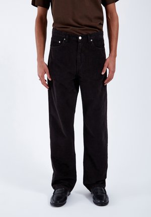 Man draagt losse zwarte corduroy broek, zwarte schoenen en een donkerbruin shirt met korte mouwen, staand tegen een effen achtergrond.