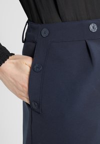 Ruka v kapsách námořnické modré sukně s dekorativními knoflíky podél bočního švu, spárované s černým dlouhým rukávem s nařaseným manžetem.