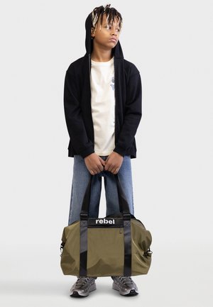 Νεαρός με dreadlocks που φοράει μαύρη μπλούζα με κουκούλα, λευκό μπλουζάκι και τζιν, κρατώντας μια χακί τσάντα ταξιδιού με την ετικέτα «rebel» μπροστά.