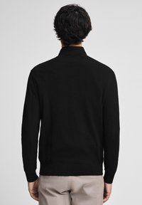 Person med kort mörkt hår iförd en svart långärmad topp och beige byxor, visad från ryggen mot en enkel bakgrund.