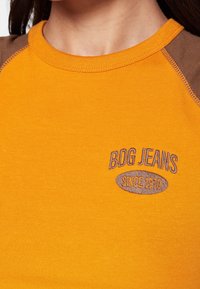Orange bomullstopp med bruna raglanärmar, med "BDG JEANS" och "SINCE 1970" broderat i brunt på bröstet. Slät textur.