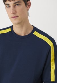 Felpa blu navy in cotone, con vivaci dettagli gialli sulle spalle e marchio. Collo arrotondato e texture morbida.