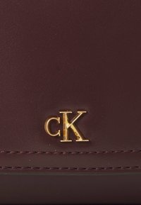Portofel din piele Burgundy, cu logo-ul auriu "cK", textură netedă și cusături contrastante pe margine. Design minimalist și formă compactă.