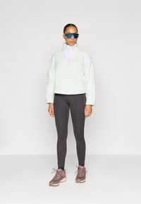 Világoszöld bolyhos pulóver negyed cipzárral és pasztell díszítéssel, fekete leggings-szel és barna sportcipővel párosítva.