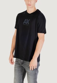 T-shirt noir à manches courtes en coton, avec un logo "AX" mat et le texte "ARMANI EXCHANGE" en gris foncé, avec une coupe décontractée.