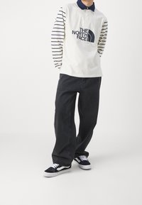 Polo tričko s dlouhým rukávem, pruhovanými rukávy, krémovým tělem a modrým límečkem; má velké grafické logo. Kombinováno s volnými černými džíny a teniskami.