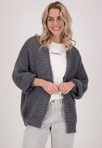 Overmaatse grijze gebreide cardigan met een gestructureerd patroon, open voorkant en losse mouwen, gedragen over een wit shirt met marineblauwe letters.