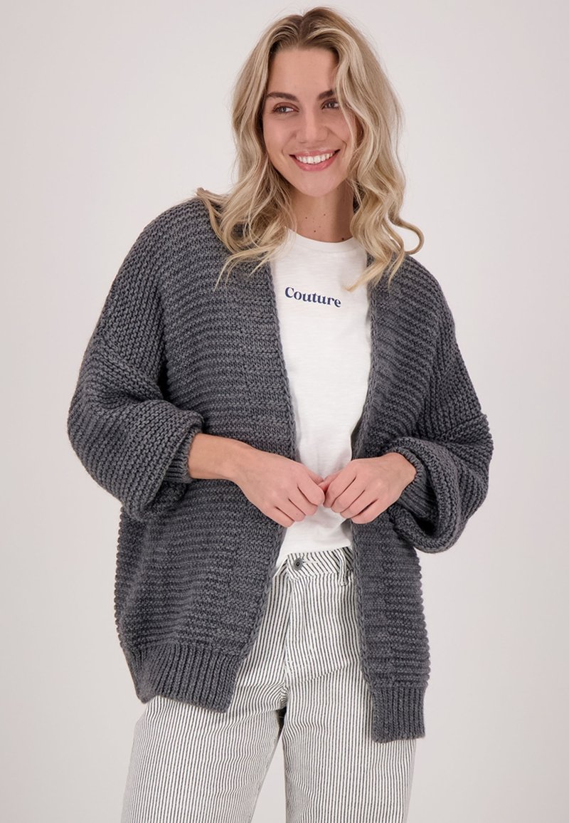 Overmaatse grijze gebreide cardigan met een gestructureerd patroon, open voorkant en losse mouwen, gedragen over een wit shirt met marineblauwe letters.