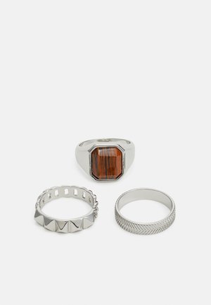 Trois bagues en argent : une avec une pierre brune rectangulaire, une avec un design en forme de coques, et une avec un motif en chevrons.