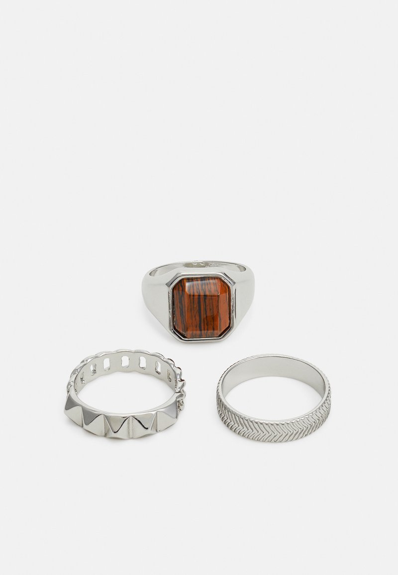 Trois bagues en argent : une avec une pierre brune rectangulaire, une avec un design en forme de coques, et une avec un motif en chevrons.