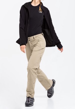 Personne en pantalon cargo beige, chaussures de randonnée noires, t-shirt noir et veste noire, marchant avec une jambe croisée devant l'autre.