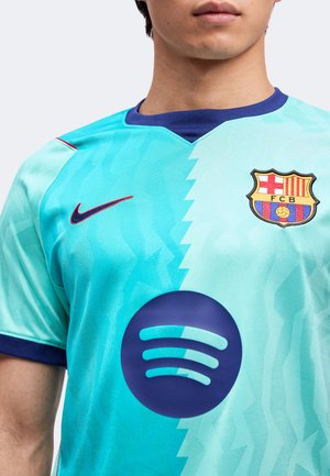 Мъж, носещ синьо и тюркоазено футболно фланелка на ФК Барселона с лого на Nike и спонсор Spotify на гърдите.