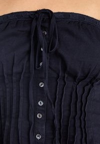 Ναυτικό μπλε αμάνικο τοπ από υφασμένο ύφασμα, με κάθετες πτυχώσεις, κουμπιά στο μπροστινό μέρος με μεταλλικά κουμπιά και λαιμόκοψη με δαντελένιο φινίρισμα.