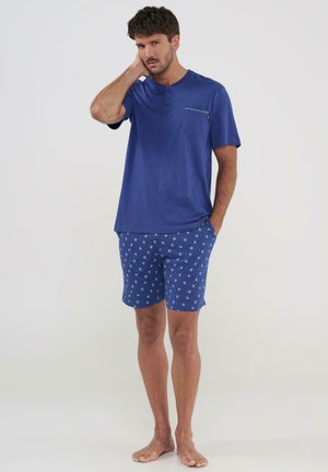 Hombre con los pies descalzos, vistiendo una camiseta Henley azul de manga corta y pantalones cortos azules con un patrón floral verde, de pie con una mano en el cuello.