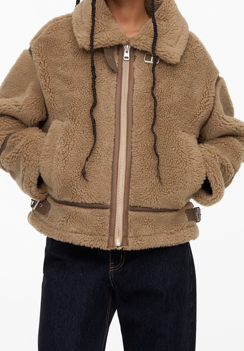 Veste en fausse peau de mouton marron avec une fermeture éclair à l'avant, un col et un ourlet ajustable. Présente des accents marron et un aspect texturé et doux.