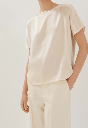 Person iført en løs, kortærmet cremefarvet bluse og matchende højtaljede bukser, med den ene hånd i lommen, foran en ensfarvet lys baggrund.