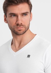 Witte katoenen V-hals t-shirt met een zwart logo op de linkerkant van de borst. De stof lijkt glad te zijn met een klassieke pasvorm.