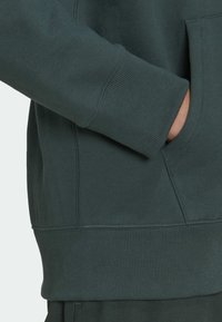 Hand halb i fickan på mörkgrön sweatshirt med ribbad mudd och matchande byxor, som visar sidesöm och sömdetaljer.