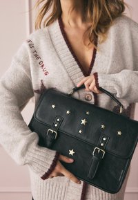 Czarna skórzana torebka zdobiona złotymi gwiazdkami, posiadająca podwójne paski i klapę, trzymana przez osobę ubraną w szary kardigan.